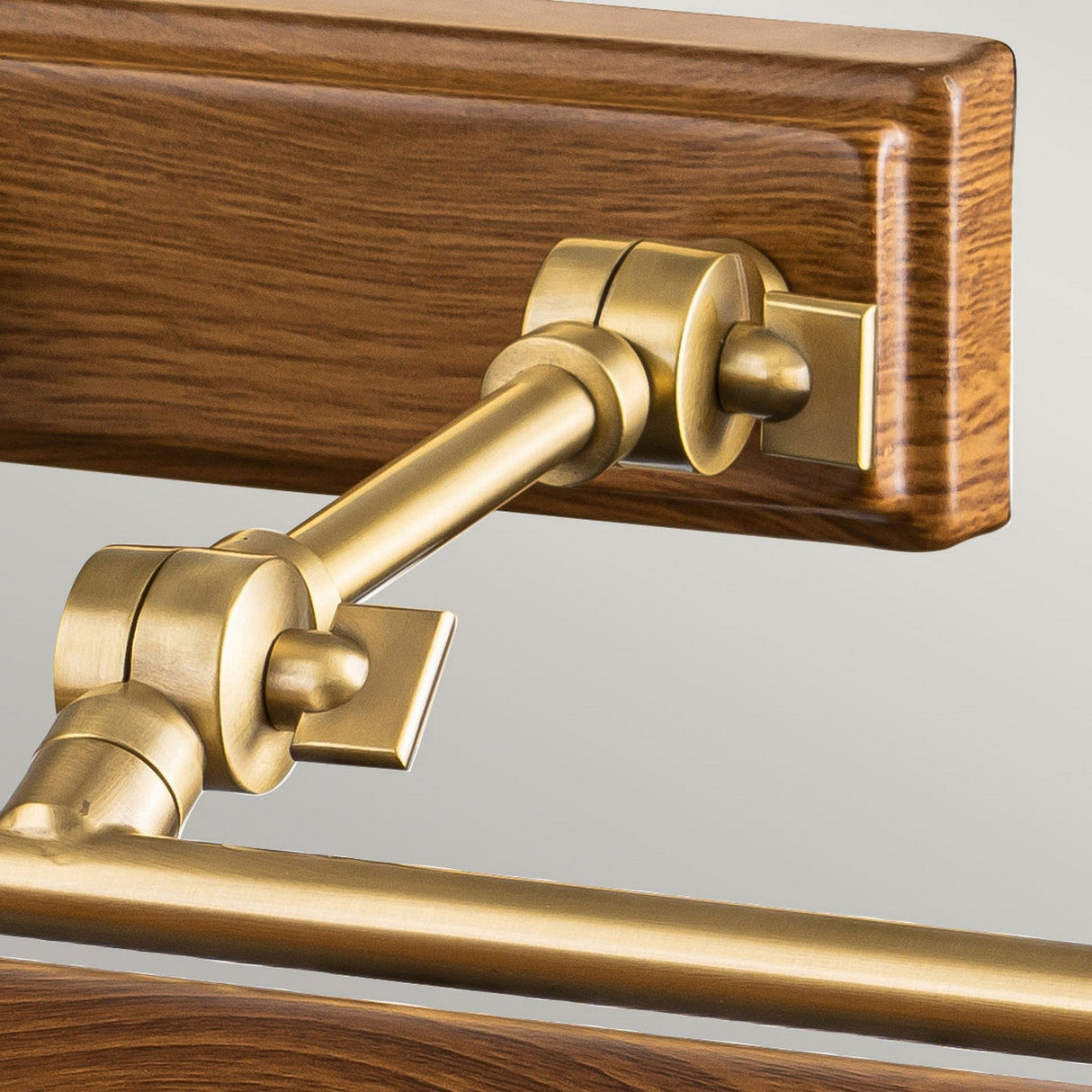 Close-up of the Winchfield 4 Light Large Picture Light, featuring an aged brass hinge mechanism attached to a wooden panel with an oak effect. The hinge, designed with multiple joints and a sleek metallic finish, connects the wood to a horizontal brass rod. This blend of materials results in a sophisticated and functional picture light design.