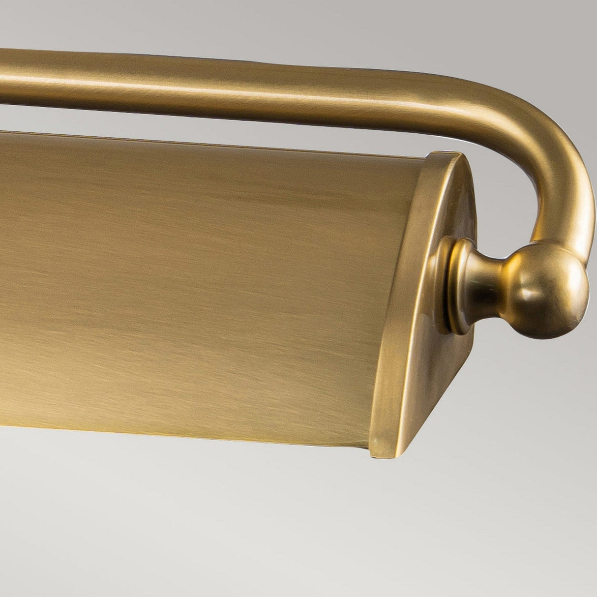 A close-up of the Winchfield 4 Light Large Picture Light in aged brass showcases its cylindrical shade and elegantly curved arm. The light's brushed finish stands out beautifully against a light gray background, reflecting its Mid Century Modern style.