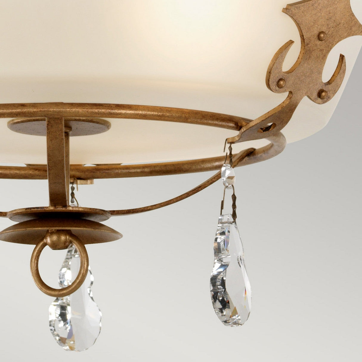 Close-up of a decorative ceiling light fixture with a frosted glass dome. It features an elegant gold patina metal frame with ornate detailing and hanging clear crystal pendants, evoking English heritage charm. The Windsor 3 Light Chandelier exudes an elegant and classic style, reminiscent of a refined chandelier.