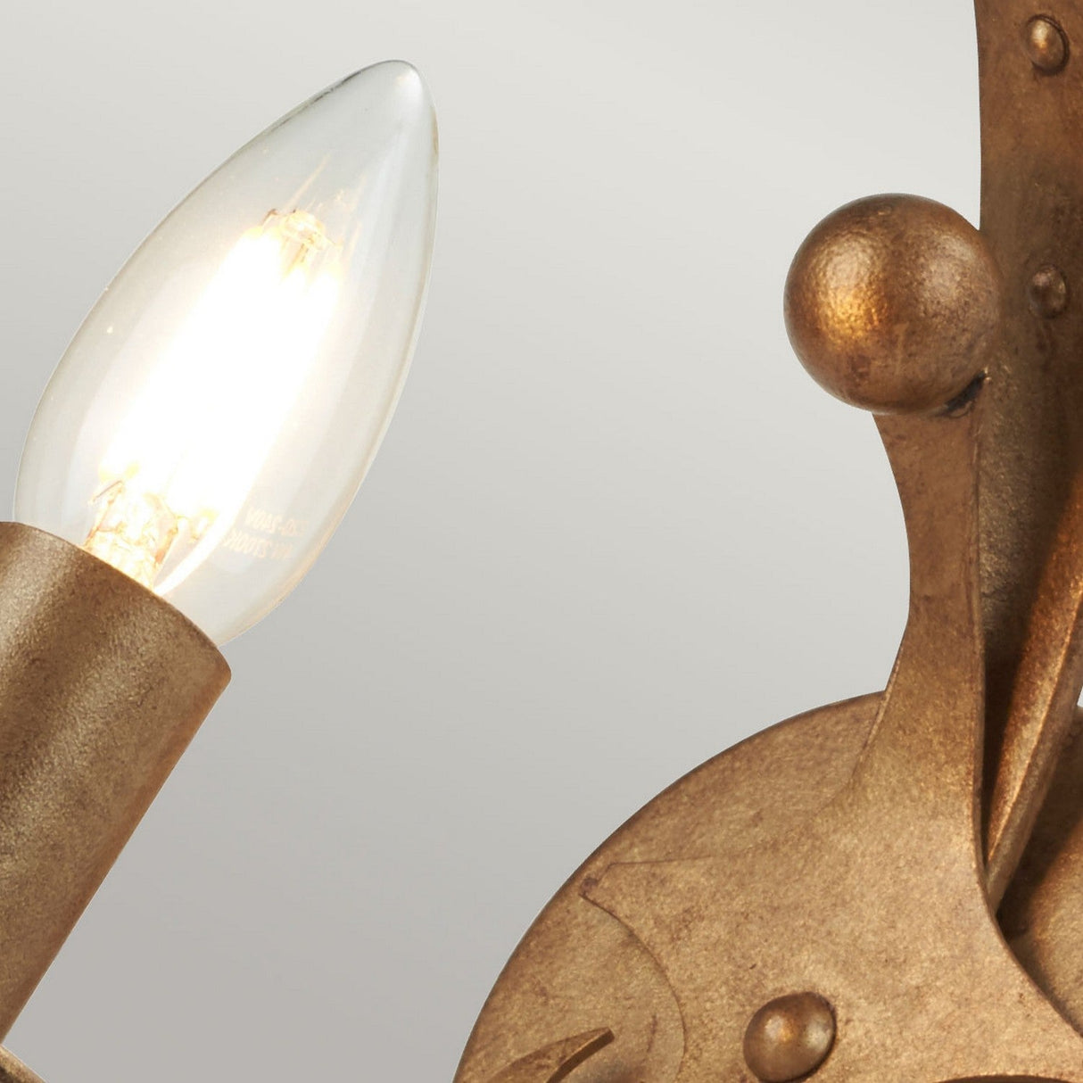 A close-up of the Windsor 1 Light Wall Light - Gold Patina showcases a lit, pointed bulb. This fixture, meticulously hand-crafted in the UK, boasts intricate details like a small decorative orb against a soft gray background.