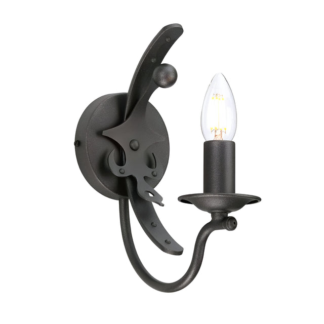 The Windsor 1 Light Wall Light - Graphite showcases a vintage-style design with a curved arm and candle-shaped light bulb. Its graphite finish and round base emphasize decorative elements inspired by British heritage, creating an antique, wrought iron look.