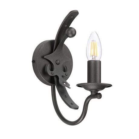 From the Windsor collection, this wall-mounted sconce features a candle-shaped bulb and winding metal arm. It is finished in dark wrought iron with a classic graphite hue, seamlessly blending British heritage with industrial elements in its circular base and curved details. The product is named "Windsor 1 Light Wall Light - Graphite.