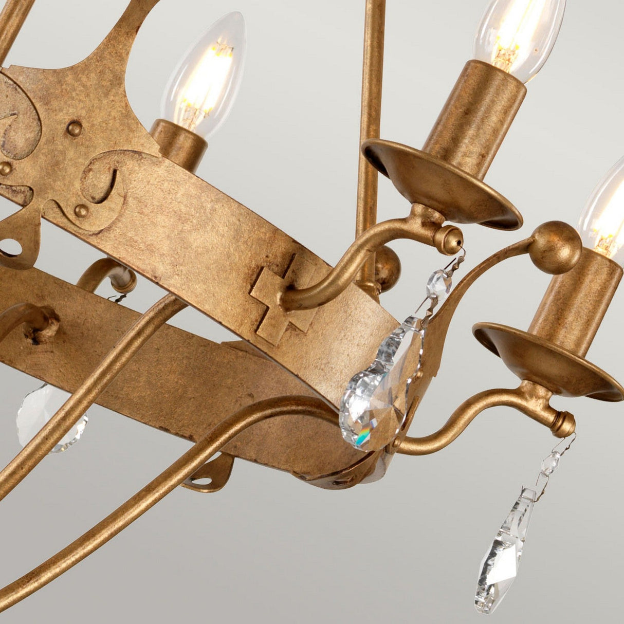 An up-close view of the Windsor 6 Light Chandelier - Gold Patina, showcasing its ornate metal arms adorned with crystal embellishments and four exposed light bulbs, set against a pale background.