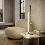 Wirmane LED Table Lamp in sand on a living room coffee table, with offset vertical bars and crossbars in aluminium and steel, a glowing white diffuser strip, beside a boucle chair.