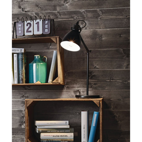 Wirnorne Table Lamp in black and white steel with bell-shaped shade and jointed arm, shown lit on a round base beside wooden crate shelves with books.