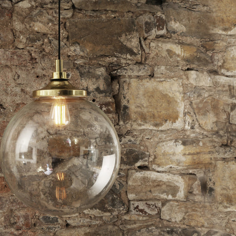 Wistella glass globe pendant light with brass gallery and stem, exposed bulb visible through clear glass, shown against a stone wall, IP44 rated.
