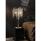The Wisteria 3 Light Table Lamp in gold with clear crystal clusters sits on a black marble pedestal beside a tufted brown velvet sofa, casting elegant dimmable lighting against a dark, leaf-patterned wall.