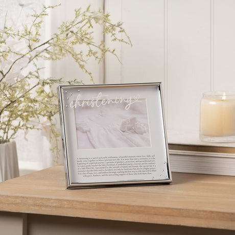 Square silver metal photo frame, 6 x 4in, with "christening" in white script and a printed verse on the mount, shown on a wooden table beside a candle and foliage.