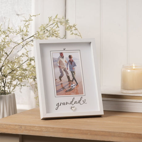 White wooden "Grandad" easel photo frame with heart detail, holding a 6 x 4in portrait, displayed on a wooden table beside greenery and a candle.