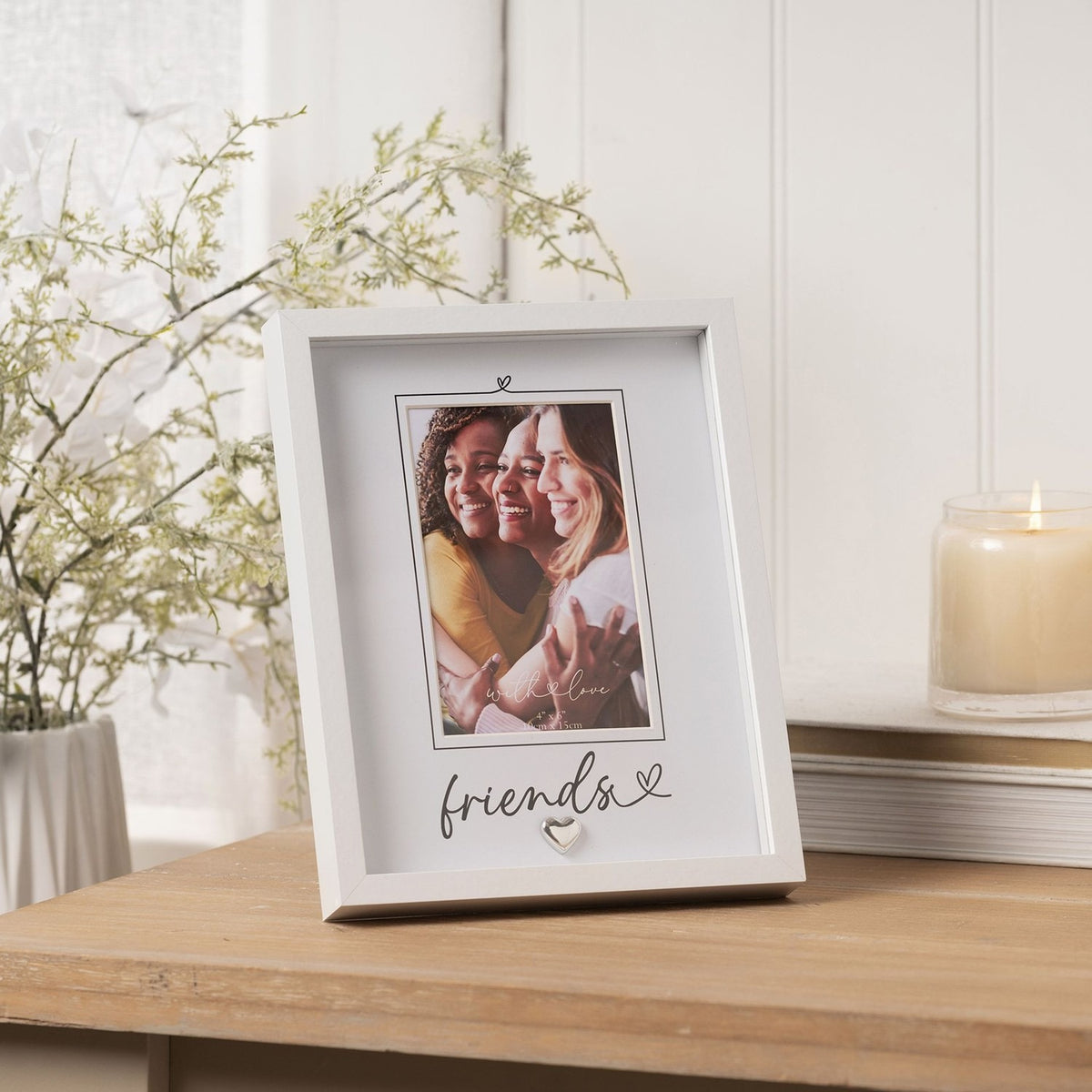 White 6 x 4in "Friends" script photo frame with silver heart detail and grey mount, shown on a wooden table beside a plant and candle.
