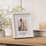 White freestanding 6x4in photo frame with "daddy" script and heart details on the mount, displayed on a wooden table beside a candle and greenery.