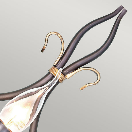 A close-up view of the Windermere 1 Light Wall Light in rust and gold highlights its decorative bulb positioned at the bottom, with intertwined designs resembling smooth, elegant tendrils that exude rustic elegance. Its wrought iron fittings beautifully contrast against the light gray backdrop.