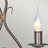 A close-up of the Windermere 2 Light Wall Light in rust and gold showcases a lit candle-shaped bulb nestled within a decorative metal holder. The bulb emits a warm glow as it's attached to a handcrafted wrought iron structure with a hook, exuding rustic charm against the plain, light gray background.