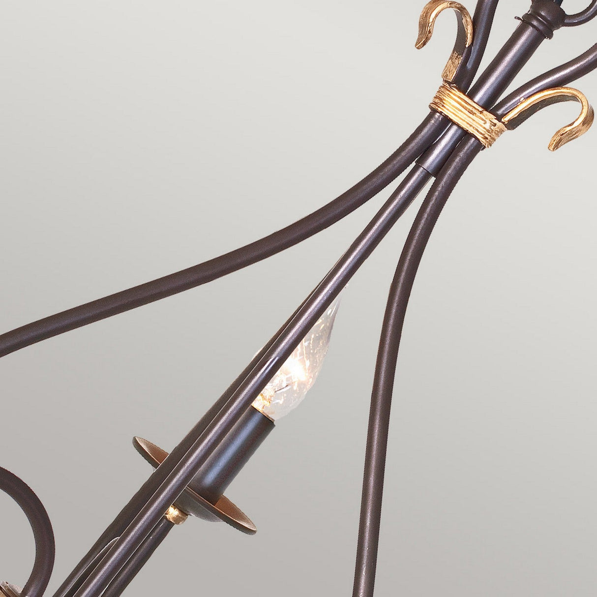 A close-up of an ornate arm from the Windermere 3 Light Chandelier in Rust/Gold, showcasing gold accents and a decorative exposed bulb. The soft gray background emphasizes the chandelier's elegant details and traditional design.