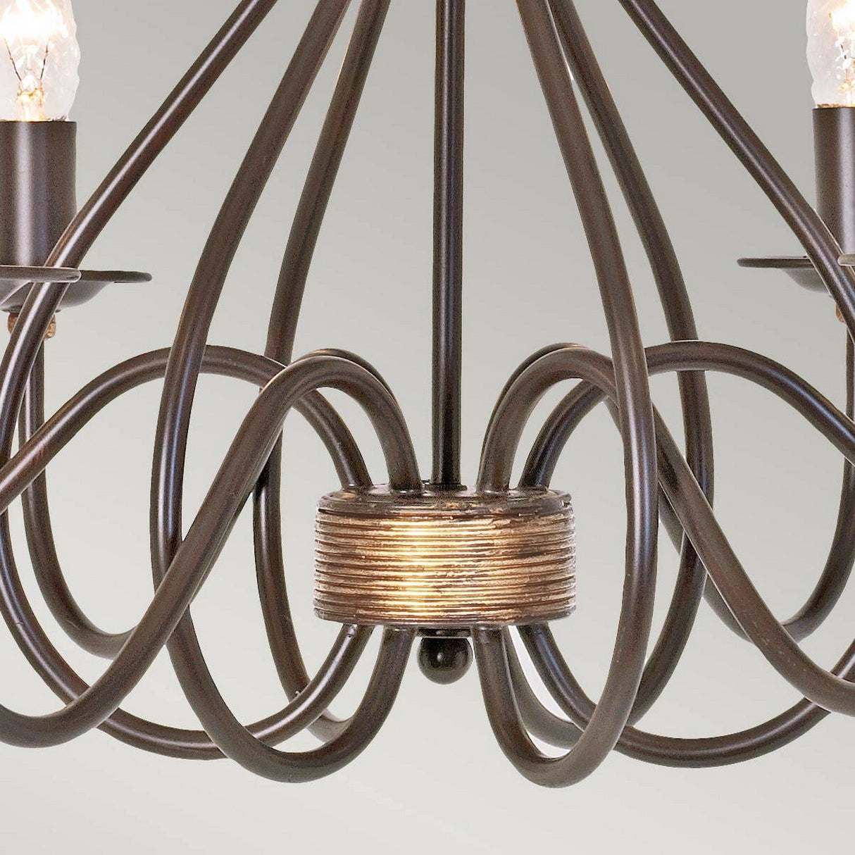 A detailed view of the Windermere 6 Light Chandelier - Rust/Gold reveals its handcrafted allure with elegant, swirling metal arms and candle-like light fixtures, perfectly highlighting its rustic charm against a light green background.