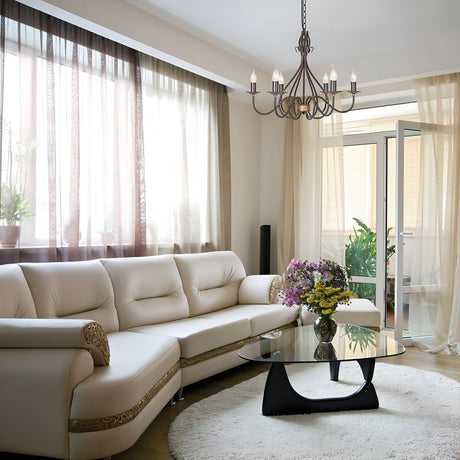 A bright living room featuring a white leather sectional sofa and a round glass coffee table adorned with a vase of colorful flowers. Large windows with sheer curtains invite natural light, while the Windermere 6 Light Chandelier in rust and gold hangs elegantly from the ceiling. Indoor plants enhance the space's charm.