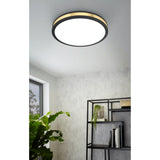 Worbette LED Flush Ceiling Light 38cm in black, round steel design with a gold band and white diffuser, shown mounted on a light grey ceiling above a styled living space with shelving and plants.