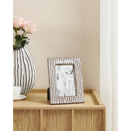 Wormiston ceramic photo frame in white and brown with ridged vertical texture, shown on a wooden sideboard beside a striped vase and pink roses.