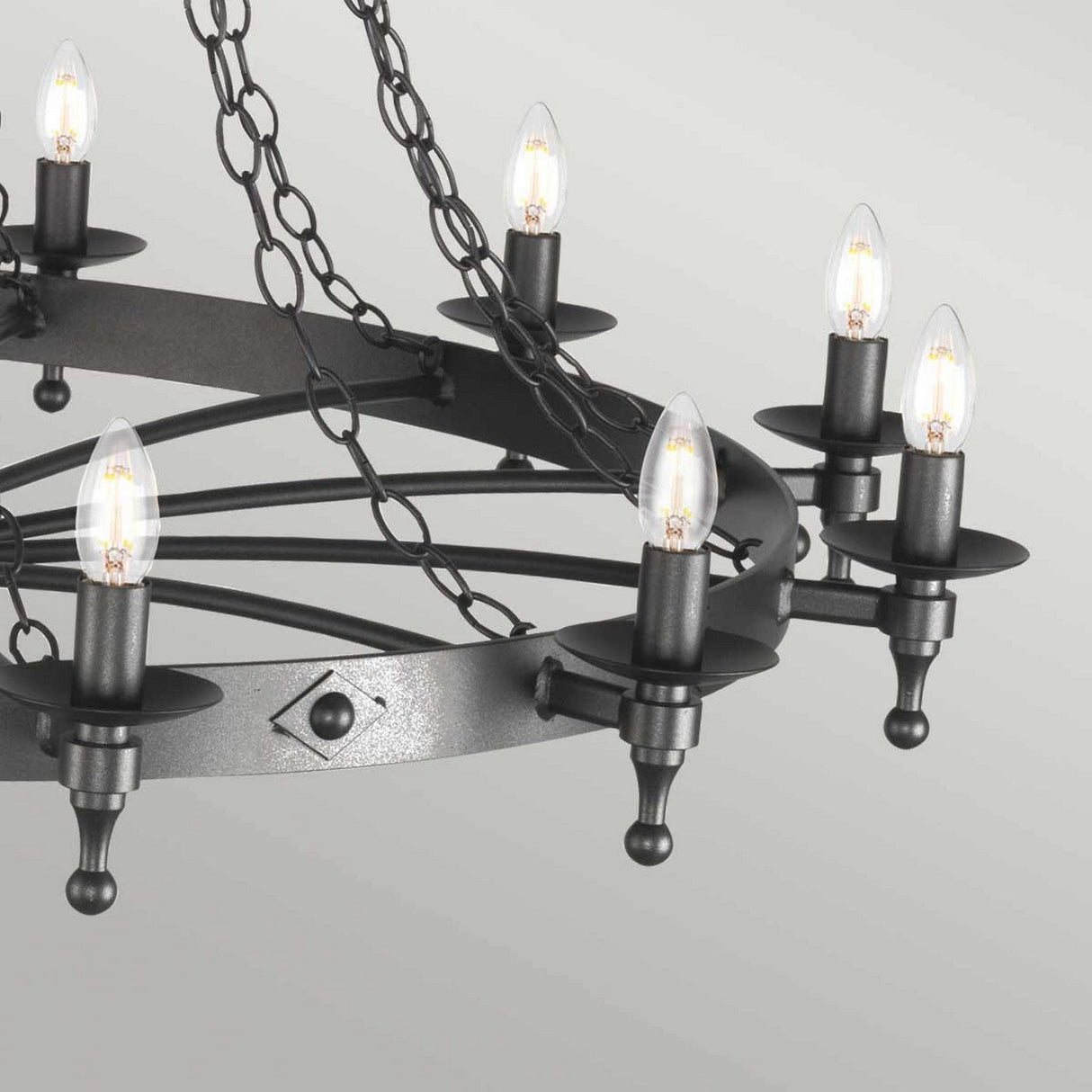 A close-up of the Warwick 18 Light Chandelier in Graphite Black showcases its medieval-inspired design, featuring a circular frame and intricate chain work complete with six candle-shaped bulbs, which stand out against the plain light grey background.