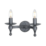 The Warwick 2 Light Wall Light - Graphite Black captures a medieval feel with its black two-light sconce, featuring candle-shaped LED bulbs and a hand-forged design. A round backplate and elegantly curved arms enhance this classic fixture.