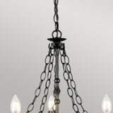 A close-up view of the Warwick 5 Light Chandelier in Graphite Black, showcasing its chain links and wrought iron-inspired frame. This fixture elegantly supports several candle-shaped bulbs, casting a gentle glow against a plain, light-colored wall.