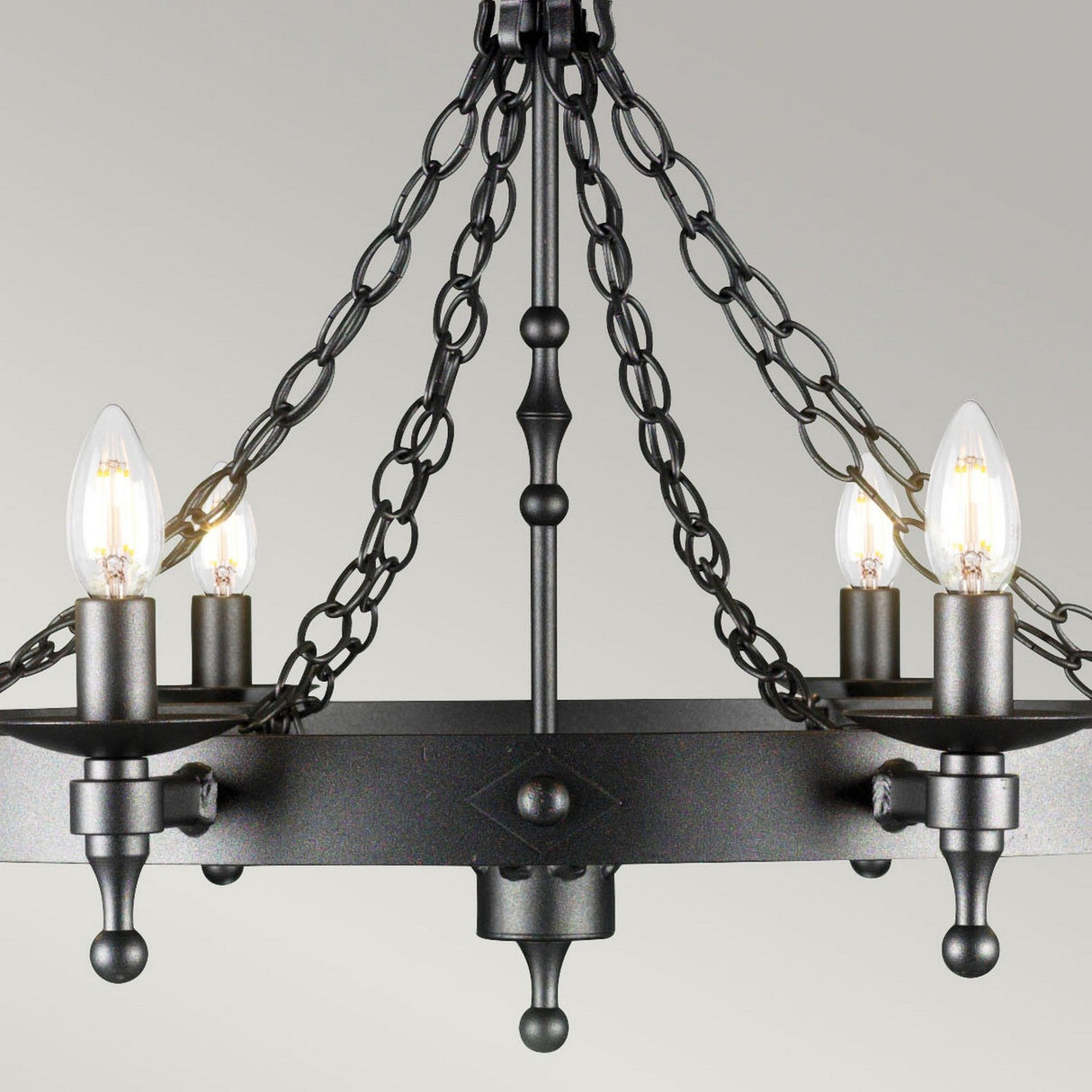 A detailed view of a graphite black wrought iron chandelier exudes rustic sophistication, adorned with four candle-shaped light bulbs. The chandelier features a circular frame held by elegant chains, skillfully combining modern and vintage elements akin to the Warwick 8 Light Chandelier in Graphite Black.