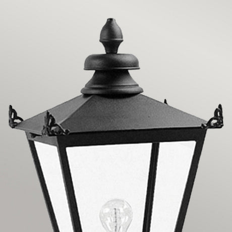 A close-up of the Wilmslow 1 Light Pedestal Light in black, designed with a vintage-style reminiscent of Victorian lighting, showcases clear glass panes that reveal a visible light bulb inside. Set against a smooth, light gray background, this elegant piece is perfect for outdoor lighting with timeless charm.