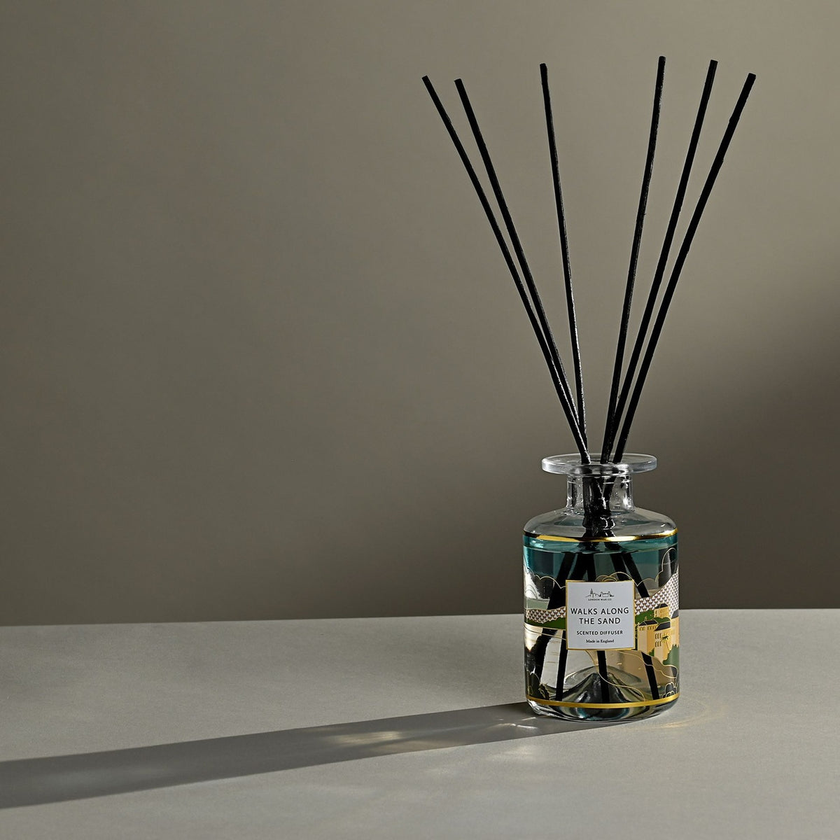 Walks Along The Sand Scented Reed Diffuser 177ml in a clear glass bottle with a teal, green and yellow landscape label, black reeds fanning upward, set against a neutral background.