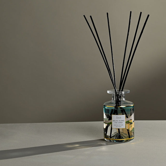 Walks Along The Sand Scented Reed Diffuser 177ml in a clear glass bottle with a teal, green and yellow landscape label, black reeds fanning upward, set against a neutral background.