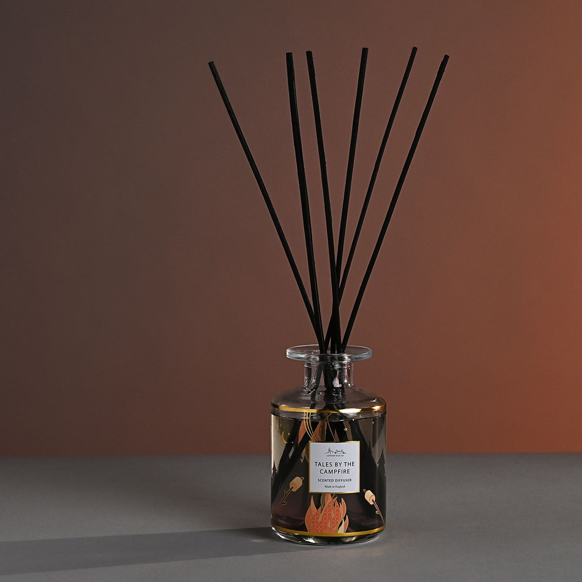 Tales by the Campfire Scented Reed Diffuser 177ml glass bottle with gold bands and campfire scene print, black reeds fanning out, set against a brown gradient background.