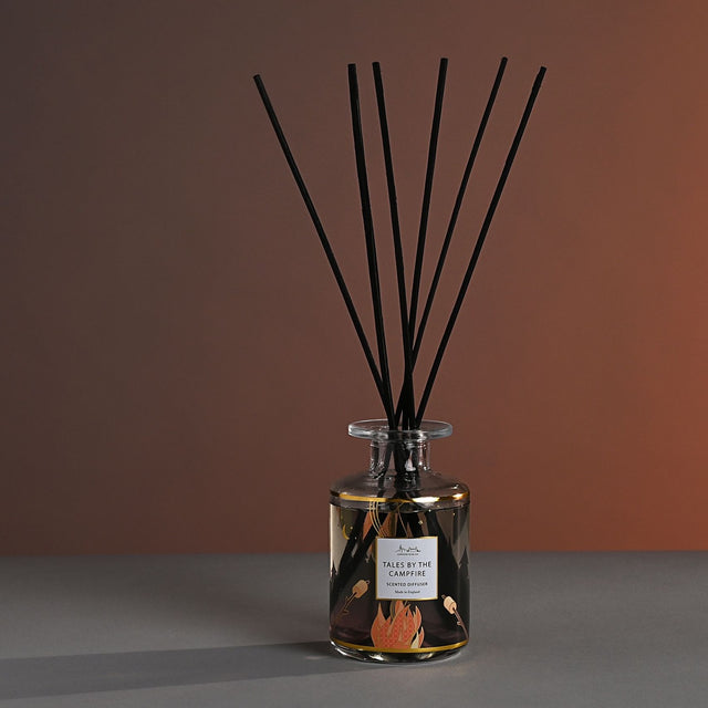 Tales by the Campfire Scented Reed Diffuser 177ml glass bottle with gold bands and campfire scene print, black reeds fanning out, set against a brown gradient background.