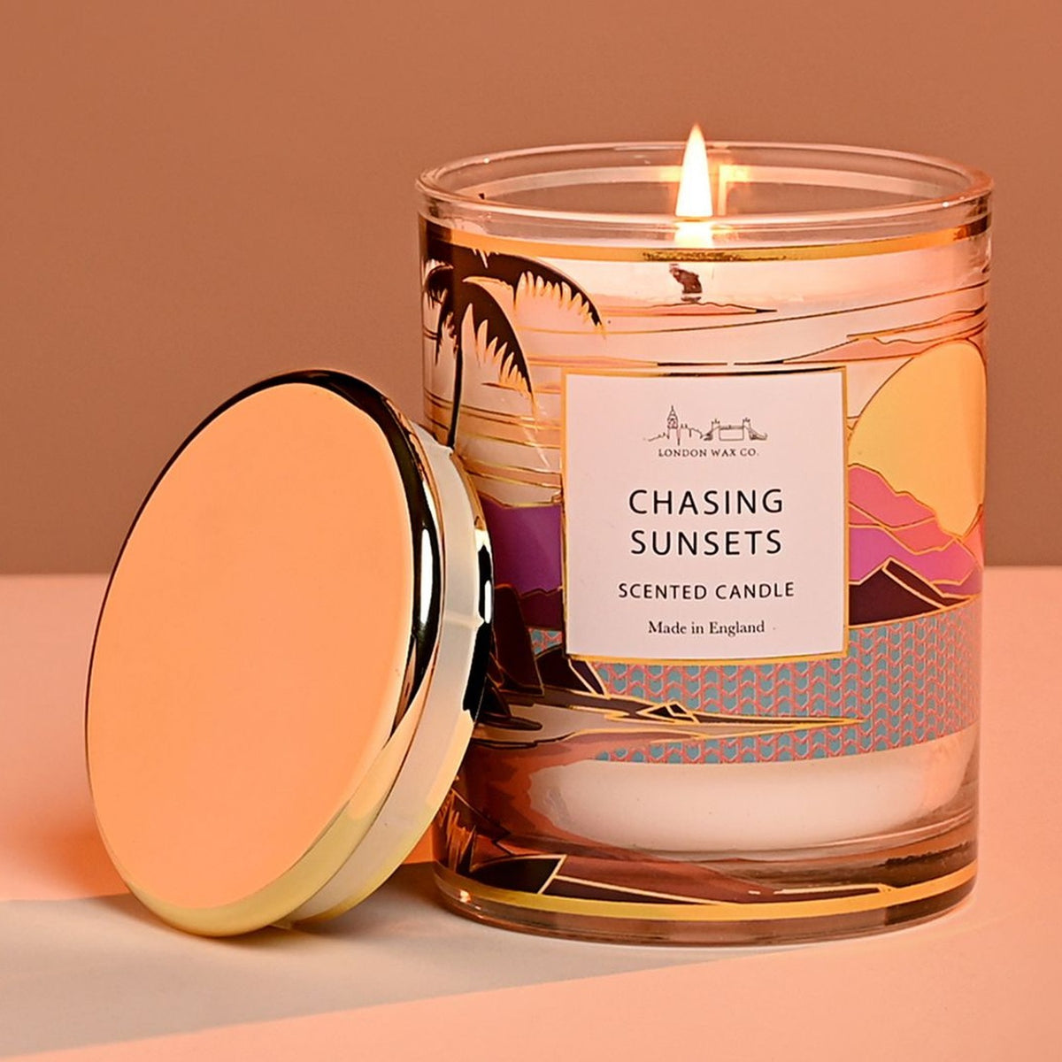 Lit single-wick Chasing Sunsets scented candle in a round glass jar with sunset, hills and palm tree design, gold rim, and matching gold lid resting beside it.
