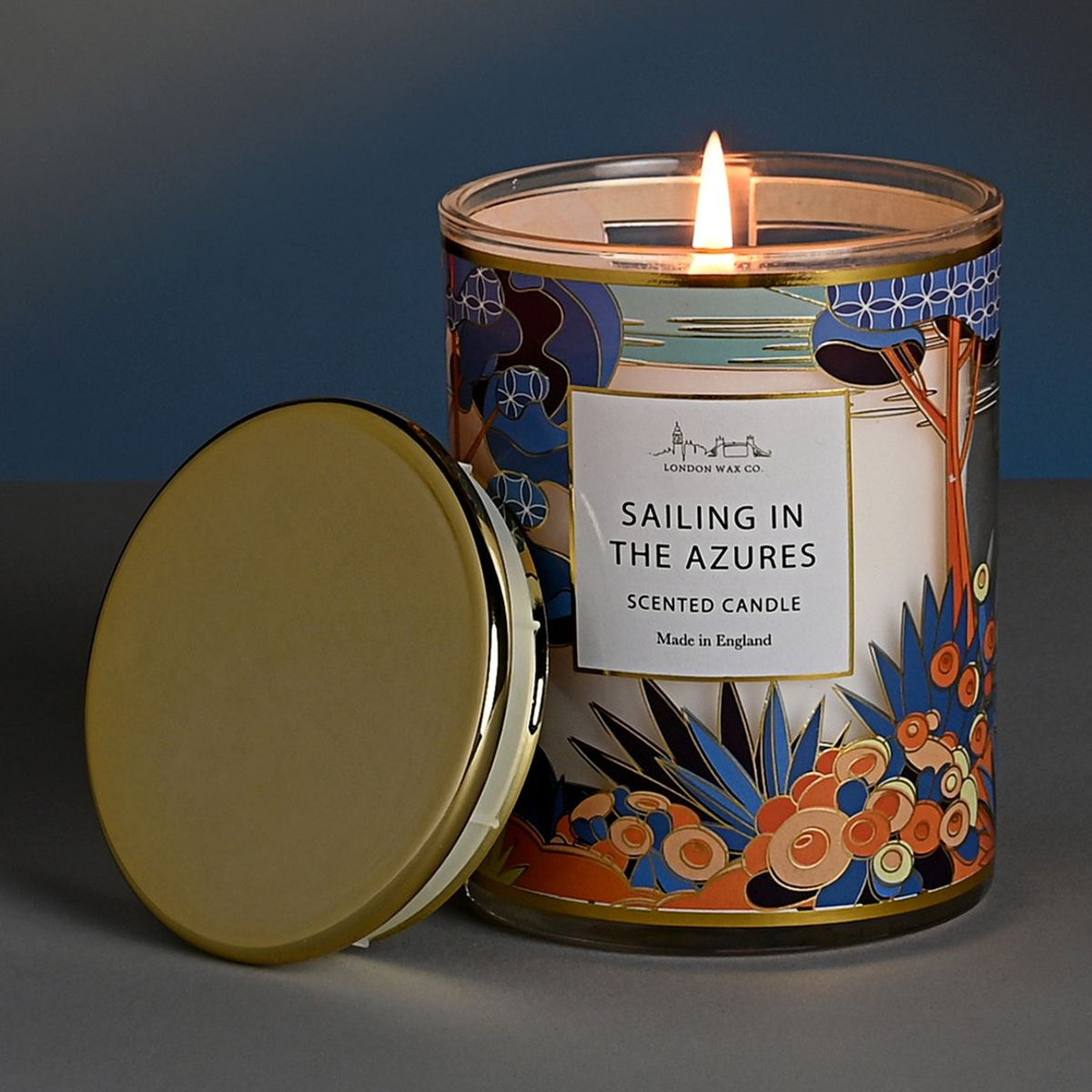 Sailing in the Azures lit scented candle in a round glass jar with floral tree and foliage design, gold rim, and matching gold lid beside it on a grey surface.