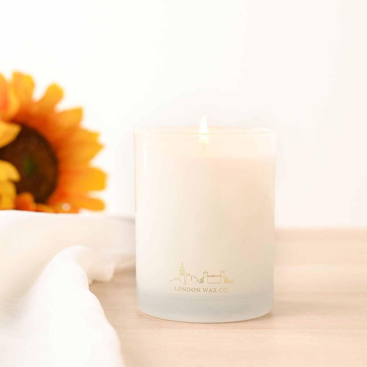 Floral Fields 1 Wick Scented Candle, lit, in a round white glass with gold London skyline design, 227g, shown with a sunflower on a light wood surface.