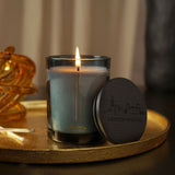 Lit round scented candle in a blue glass vessel with gold London skyline design, shown beside its black lid featuring the same skyline engraving, on a gold tray.