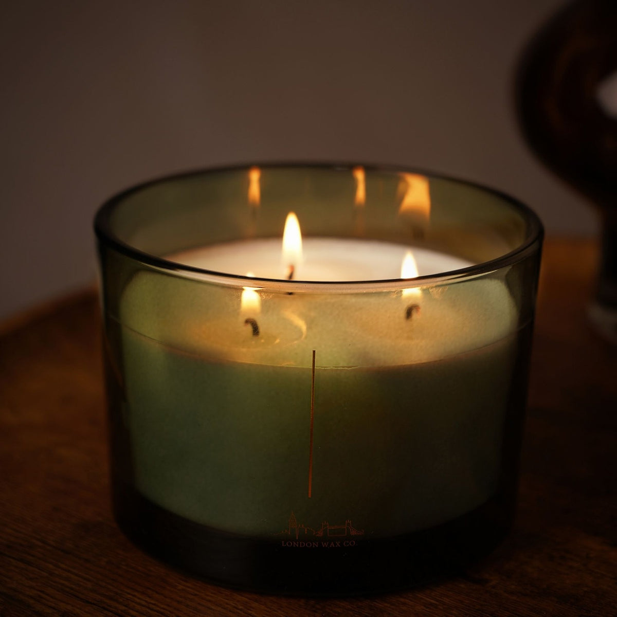 Enjoy the Silence Round 3 Wick Scented Candle 397g in Green, lit with three flames in a smoky green glass vessel, gold skyline design on front, dark wood surface.