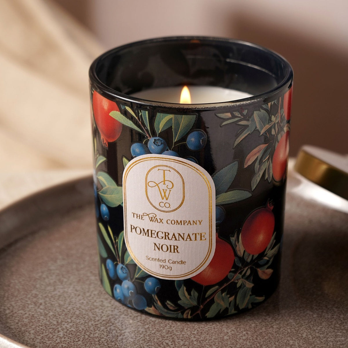Pomegranate Noir scented candle in a round black glass jar with pomegranate, blueberry and leaf print, lit wick visible, shown beside a gold lid on a tray.