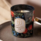 Pomegranate Noir scented candle in a round black glass jar with pomegranate, blueberry and leaf print, lit wick visible, shown beside a gold lid on a tray.