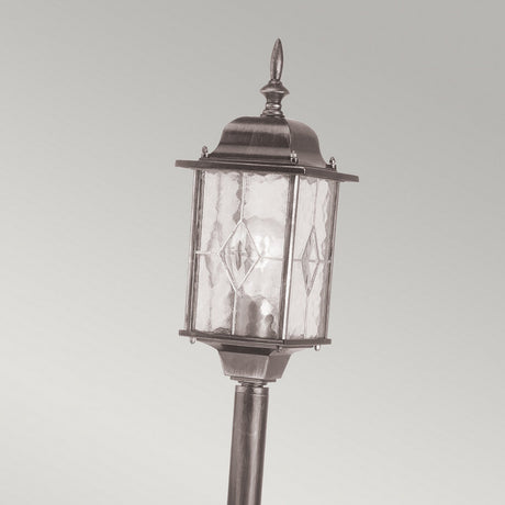 Introducing the Wexford 1 Light Pillar Light in Black/Silver: This vintage-style outdoor lamp is crafted from durable materials and showcases a clear glass lantern that encases a light bulb. It features decorative metal elements and a pointed top, embodying traditional design against a plain, light gray background.