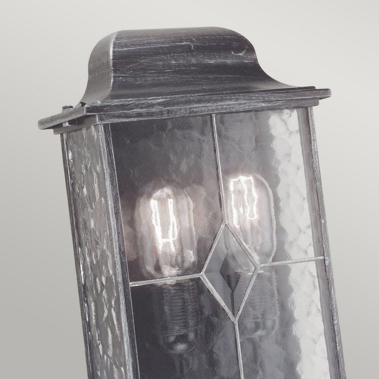A detailed view of the Wexford 2 Light Outdoor Wall Light - Black/Silver, highlighting its vintage design with hand-leaded glass panes and an elegant diamond-shaped embellishment. This outdoor wall lantern showcases two Edison bulbs, softly illuminating a neutral backdrop and embodying traditional sophistication.