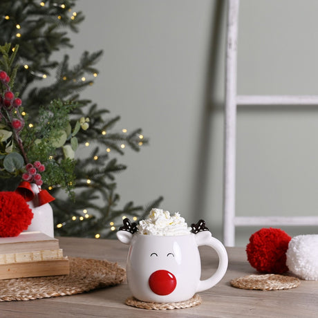 White stoneware Reindeer Mug with raised red nose, antler ears, and closed smiling eyes, filled with whipped cream, styled on a festive table with Christmas décor.