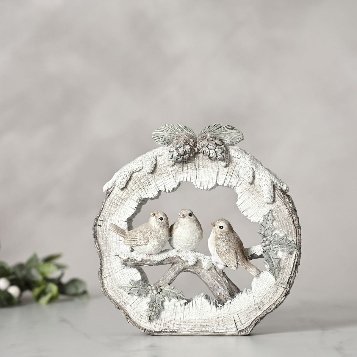 Round log resin figurine in white, featuring three robins perched on a snowy branch with pinecones and holly, set against a plain light grey background.
