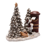 Resin Christmas figurine of a boy in a white coat and red scarf posting a letter into a tall red pillar box, beside frosted fir trees and a gold sleigh with parcels on a snowy base, white background.