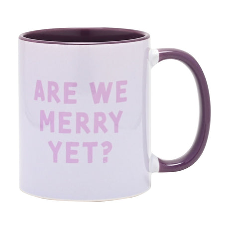 White stoneware mug with purple interior and handle, "ARE WE MERRY YET?" printed in lilac-pink capitals on the white body, 11oz, plain background.