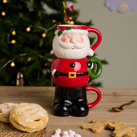 Set of 3 Christmas Santa stacking glazed stoneware mugs in red, forming his face, jacket and black boots, shown on a wooden table with mince pies and gingerbread near a Christmas tree.