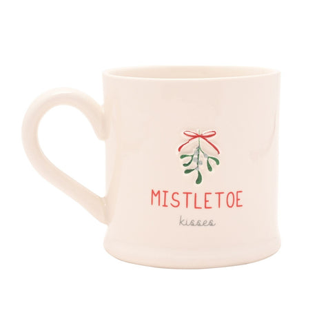 White stoneware mug with embossed mistletoe sprig and red bow, "MISTLETOE kisses" printed in red capitals and grey script, shown on a plain white background.
