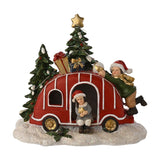 Light-up Christmas Caravan decoration in red plastic with white trim and gold wheels, roof piled with gifts, two Santa-hatted children figures, snowy trees, white background.