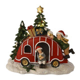 Light-up red Christmas Caravan decoration with gift boxes and gold star on roof, two children in Santa hats beside snow-dusted trees, on white background.