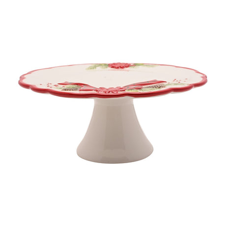 Round white ceramic cake stand on a flared footed base, with a scalloped red-edged rim and raised poinsettia, pine, and tartan bow decoration, shown on a plain white background.