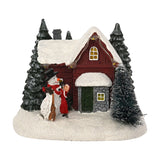 Light-up plastic Christmas decoration of a red-walled, snow-roofed house with fir trees, a top-hatted snowman, and a red-coated figure, on a plain white background.